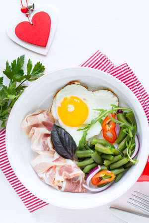 Breakfast for your beloved for the holiday: heart-shaped egg, bacon, green beans on a white table. Selective focus. View from above. Copy spaceの写真素材