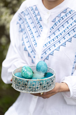 young girl in a white embroidered dress holds a basket with colored Easter eggs in her hands....