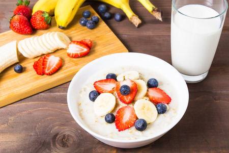 Oatmeal with banana, strawberry and blueberry and glass of milk on brown wooden tableの写真素材