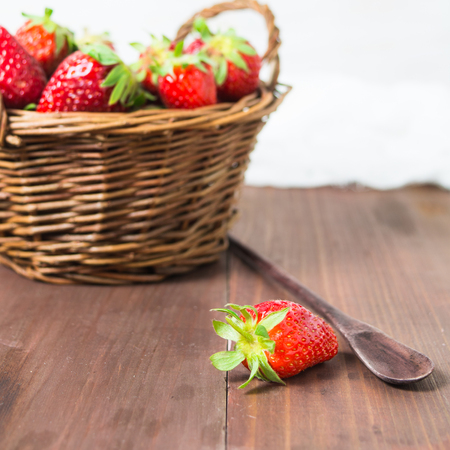 One strawberry on foreground and basket on background over brown wooden table, square shotの写真素材