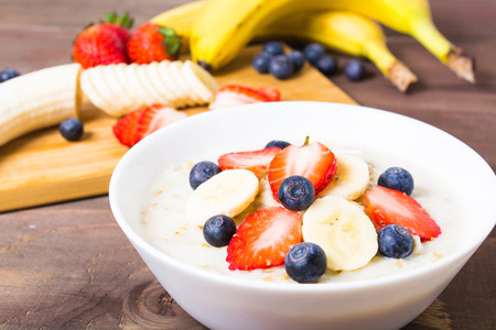 Milk oatmeal decorated with fruits (banana, strawberry, blueberry, milk) closeup on brown wooden tableの写真素材