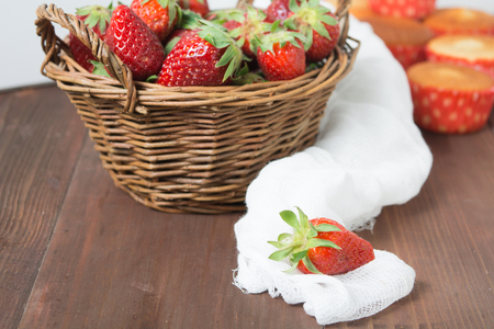 Basket of strawberry on brown wooden table and one strawberry in focus with some muffins on backgroundの写真素材