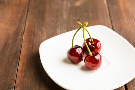 Three cherry on white plate on brown wooden backgroundの写真素材