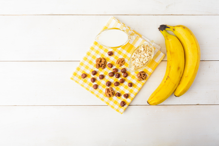 Ingredients for healthy breakfast with fresh bananas, nuts and oatmeal with milk over white wooden backgroundの写真素材