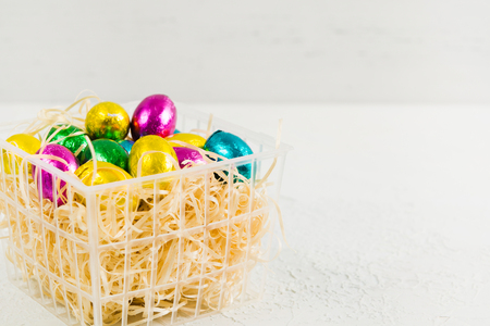 Colorful Easter chocolate eggs in basket with hay on white background with copy space, selective focusの写真素材