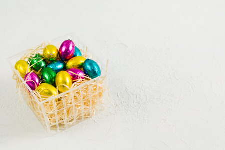 Chocolate candies easter eggs in plastic basket with copy space on white textured background