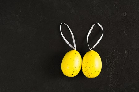 Easter concept with egg decoration. Two yellow colored eggs with dots with ribbons over black textured concrete background. Flat lay. Top viewの写真素材