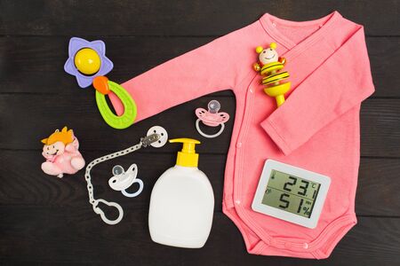 Baby accessories and digital thermo-hygrograph on dark wooden background. Pink bodysuit, two rattles, two pacifiers, toy and soap with hygrometer and thermometer for baby health. Baby shower. Flat lay. Top viewの写真素材