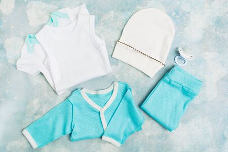 Newborn baby clothes - white bodysuit, blue shirt, blue pants, white hat and pacifier on blue textured background. Baby shower concept for partyの写真素材