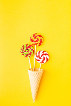 Three colorful candies on stick in waffle cone over yellow background with copy space. Top view. Flat lay. Vertical shotの写真素材