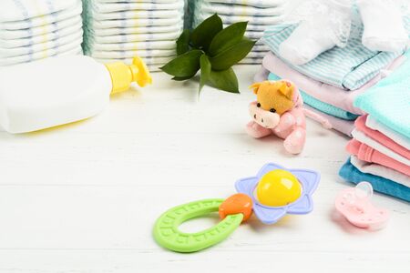 Baby accessories with shower gel or soap, pacifier, rattle, clothes and diapers over white wooden background. Set for newborn with copy spaceの写真素材