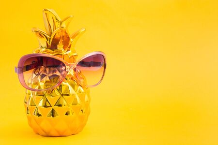 Golden pineapple decoration with pink sunglasses on yellow beach background with copy space. Summer vacation concept with vitamins and sunの写真素材