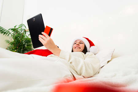 Caucasian woman in Santa hat lying on bad with tablet and plastic cardの写真素材