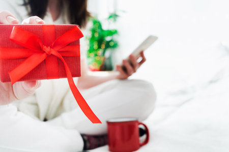 Faceless woman sitting on bed at home with phone and showing red gift with bowの写真素材