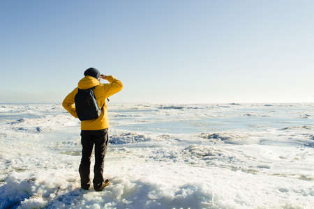Faceless man in yellow raincoat with backpack standing and looking on frozen seaの写真素材