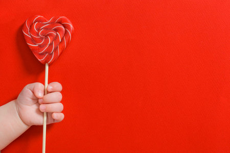Newborn baby hand holding sweet candy heart on stick on red backgroundの写真素材