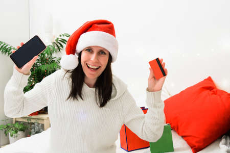 Beautiful smiling woman in Santa hat holding phone and bank plastic cardの写真素材