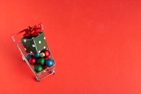 Shopping cart on wheels with Christmas ornaments and gift box on red backgroundの写真素材
