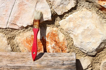 Old red paint brush drying on piece of wood, leaning on stone wall in the sunlightの写真素材