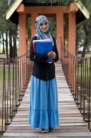 beautiful young muslimah woman with head scarf holding the books and standing on hanging bridge at recreation parkの写真素材