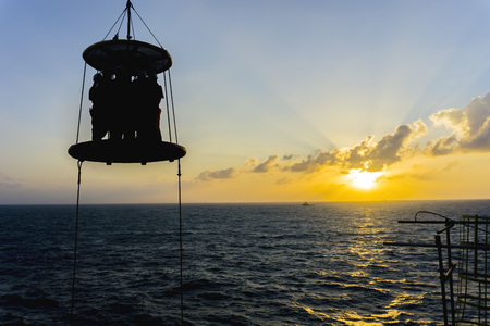 Silhouete people.A group of offshore crew crossing the sea from barge to oil and gas platform using transfer basket during sunrise with rays of light.の写真素材