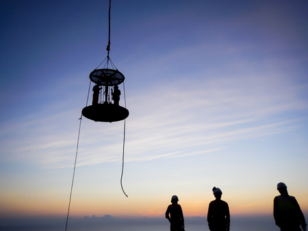 Oil and gas industry. Silhouette of offshore workers standing on helideck area to use Personal Transfer Basket (PTB) for transferring from oil and gas platform to Accommodation Work Barge (AWB) during sunsetの写真素材