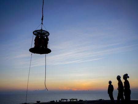 Oil and gas industry. Silhouette of offshore workers standing on helideck area to use Personal Transfer Basket (PTB) for transferring from oil and gas platform to Accommodation Work Barge (AWB) during sunsetの写真素材