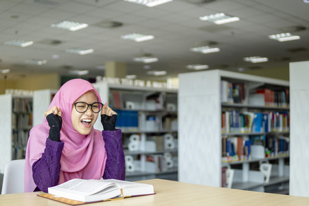 A beautiful Muslimah student sitting in the library with book open on the table shows exciting and happy facial expression.の写真素材
