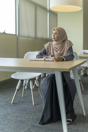 A beautiful Muslimah student sitting in the library and pensive thinking about something.の写真素材