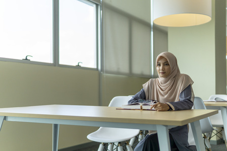 A beautiful Muslimah student sitting in the library and pensive thinking about something.の写真素材