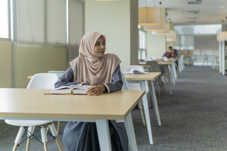 A beautiful Muslimah student sitting in the library and looking something at her left side.の写真素材