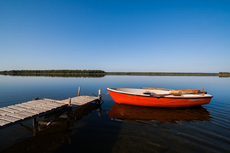 A beautiful idyllic view of a glassy lake in northern Sweden. The sky is perfectly blue and you see a rowing boat with fishing gear docked by a small jetty.の写真素材