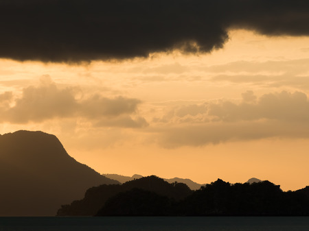 An amazing sunset view in the tropics. The sky is golden orange and the mountains in the distance look magical. Dark clouds frame the picture perfectly. Location: Tanjung Rhu Beach, Langkawi, Malaysia.の写真素材
