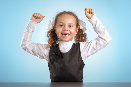 Happy Little Schoolgirl Sits At Table With Raised Hands On Blue Backgroundの写真素材