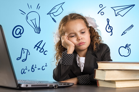 Sad Little Girl Sits At Table With Books Near Laptop On Blue Background. Back To School Themeの写真素材