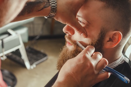 Young Bearded Man Getting Beard Haircut With A Straight Razor By Barber. Barbershop Themeの写真素材