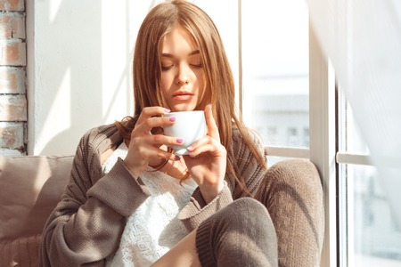 Beautiful Young Woman Sitting On A Windowsill With Cup
