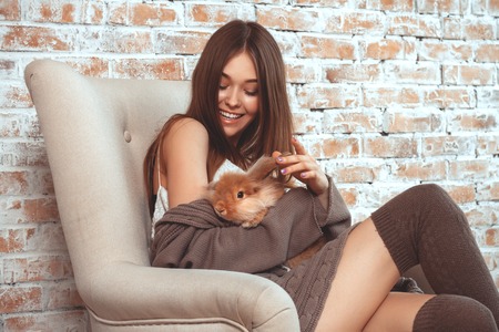Beautiful Young Woman Sitting In Armchair On Brick Wall Background And Playing With Easter Bunnyの写真素材