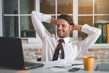 Handsome Young Businessman Sitting At His Workplace With Hands Behind Head And Looking At His Laptop. Business Themeの写真素材