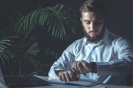Handsome Young Bearded Businessman Sitting At His Workplace And Looking At His Watch. Business Themeの写真素材