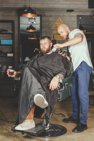Serious Bearded Man Getting Haircut By Barber And Holding A Glass Of Whiskey While Sitting In Chair At Barbershop. Barbershop Themeの写真素材