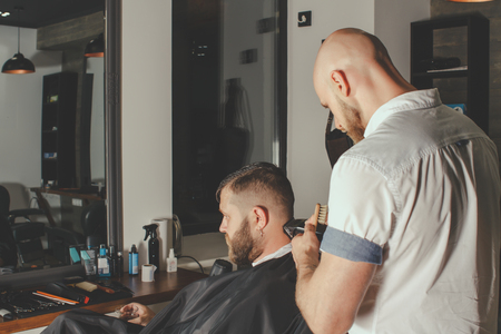 Serious Bearded Man Getting Haircut By Barber And Holding A Glass Of Whiskey While Sitting In Chair At Barbershop. Barbershop Themeの写真素材