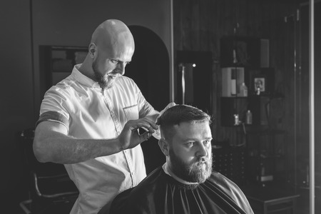 Serious Bearded Man Getting Haircut By Barber While Sitting In Chair At Barbershop. Barbershop Themeの写真素材