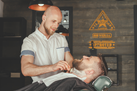 Serious Bearded Man Getting Beard Haircut By Barber While Sitting In Chair At Barbershop. Barbershop Themeの写真素材