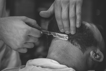 Serious Bearded Man Getting Beard Haircut With A Straight Razor By Barber While Sitting In Chair At Barbershop. Barbershop Themeの写真素材