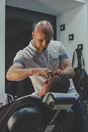 Serious Bearded Man Getting Beard Haircut By Barber While Sitting In Chair At Barbershop. Barbershop Themeの写真素材