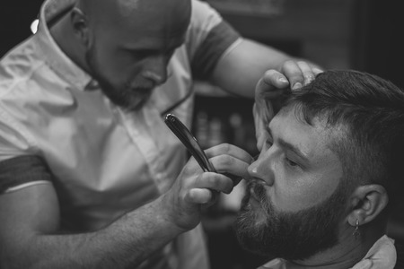 Serious Bearded Man Getting Beard Haircut With A Straight Razor By Barber While Sitting In Chair At Barbershop. Barbershop Themeの写真素材