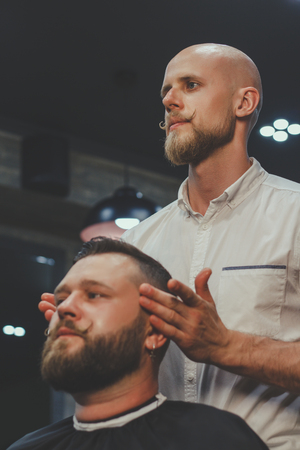 Serious Bearded Man Getting Haircut By Barber While Sitting In Chair At Barbershop. Barbershop Themeの写真素材