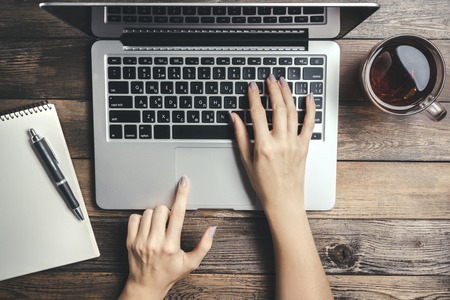 Female Hands Typing On The Keyboard Of A Laptop On Vintage Wooden Table. Mock-up With Laptop, Cup And Pan On Notebookの写真素材