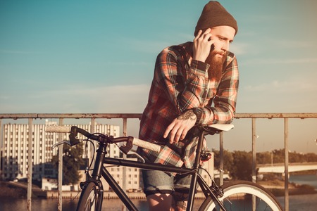 Handsome Bearded Man In Red Shirt Stops Cycling, To Speak On His Phone On A Bridge Near The Riverの写真素材